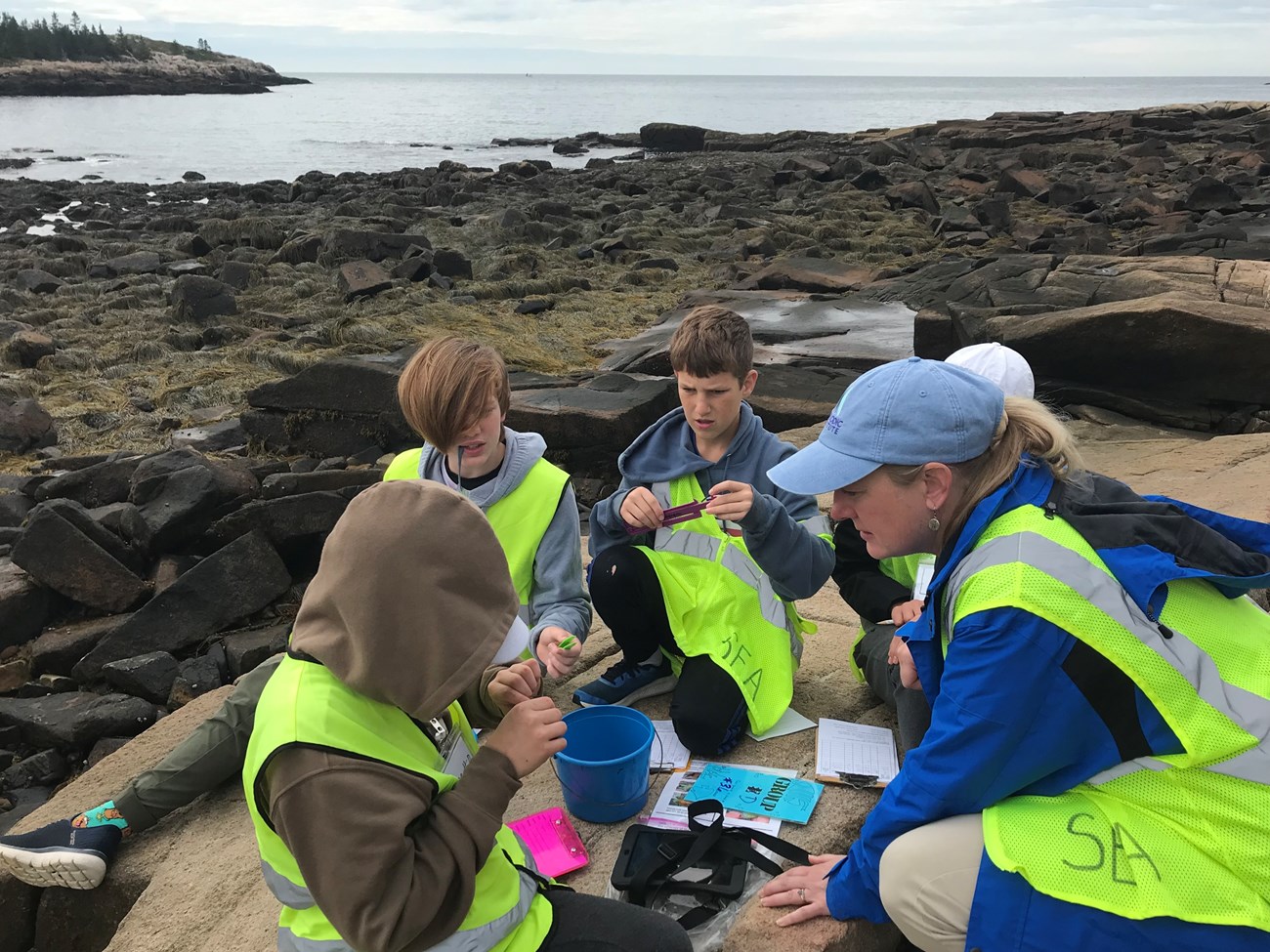 A woman and four teenagers in bright vests sit on a large rock by the ocean. The teenagers are holding plastic measuring devices and the woman is watching closely.