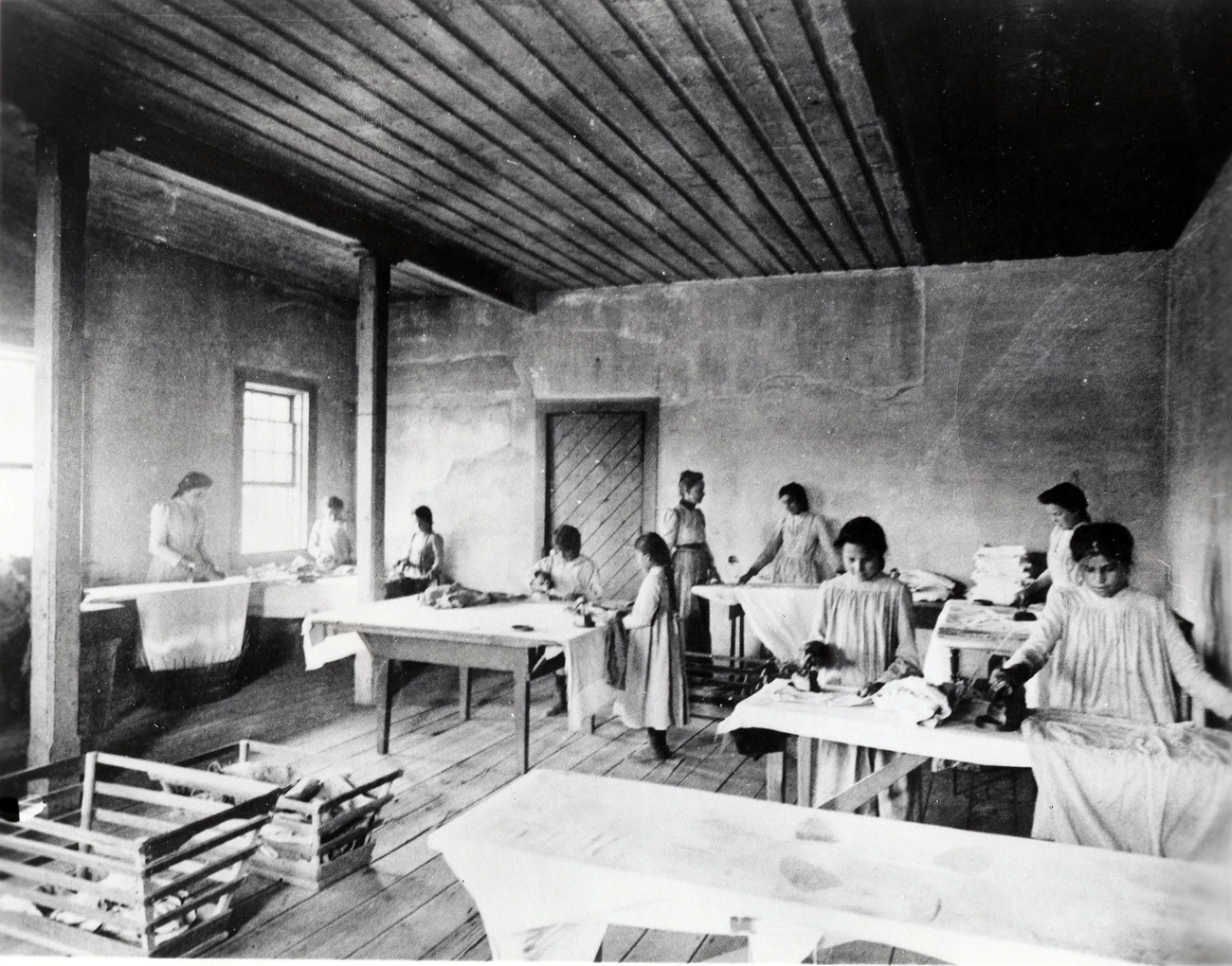 Nine Indigenous girls in western-style dresses and haircuts iron laundry as white teacher looks on.