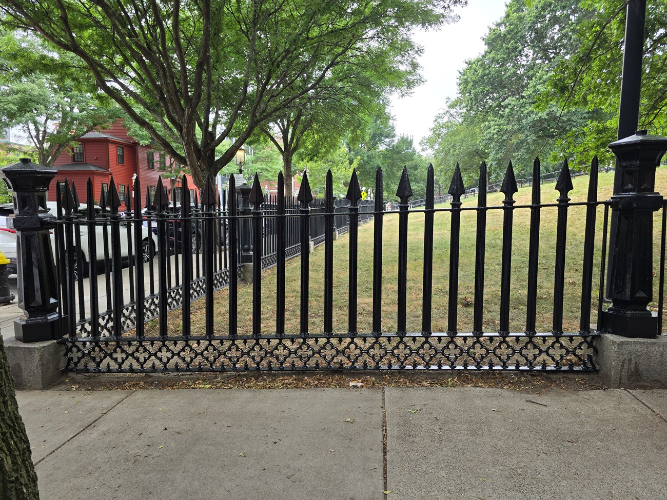 Section of black wrought-iron fence. At each end is a decorative pillar, and there are 22 bars between the pillars. The tip of each bar alternates between a straight spike or triangular spike. The bottom section of the fence has a floral design.