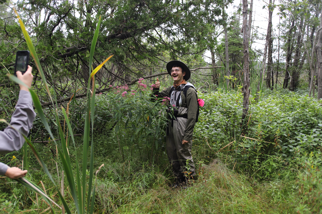 A park ranger smiles and gives a thumbs up. He is standing next to a tall green plant with pink flowers, which comes up to his chin. Another ranger is taking his picture.