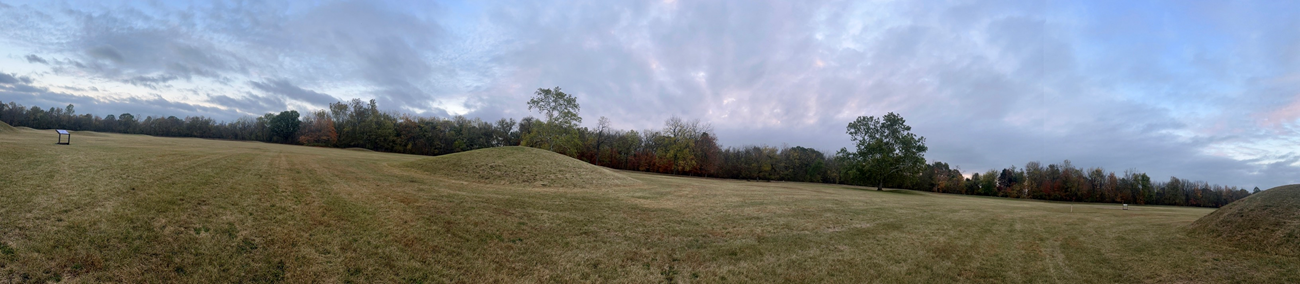 Earthen mounds in a large grassy area surrounded by trees with autumn leaves.