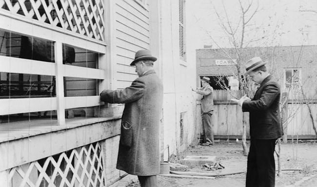 Two men measure a building as another stands by taking notes.