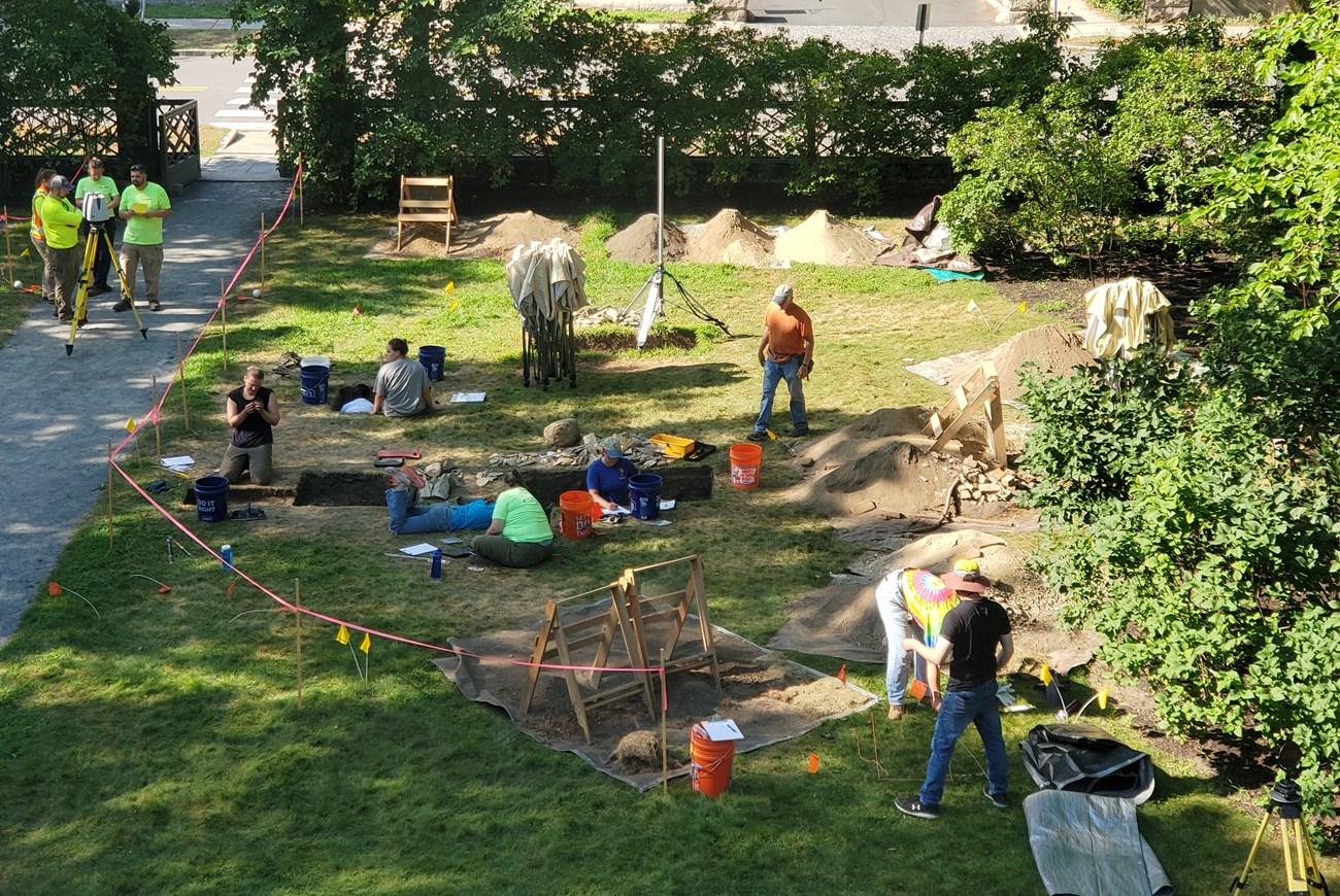 11 people stand in and around rectangular holes in a lawn with piles of dirt