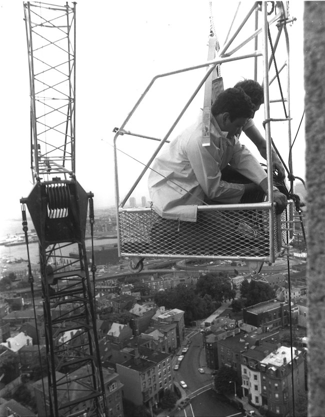 A crane holding a small metal work basket beside the monument