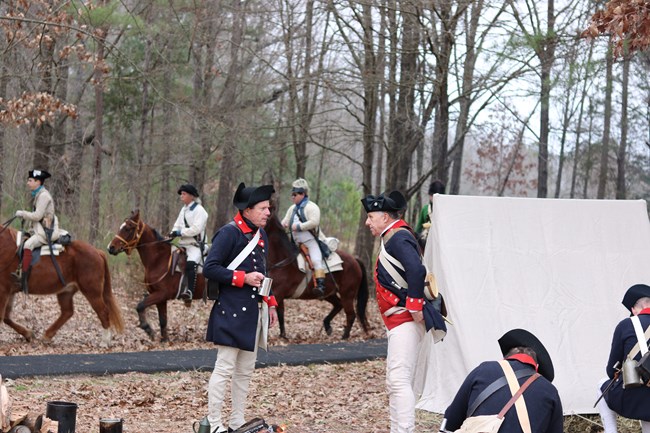 Two reenactors speaking while reenactors ride behind them on horseback