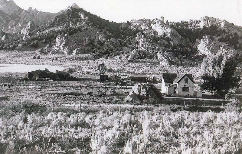 Rustic stone farmhouse sits in field surrounded by arid landscape and rocky mountains.