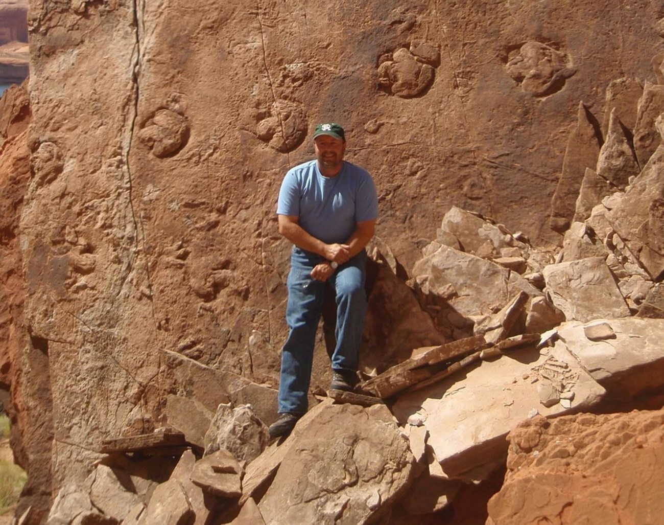 a man with a beard and ball cap stands next to a large boulder with a track of dinosaur footprints clearly visible