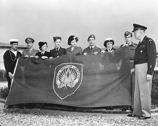 A black and white image showing General Eisenhower and a row of people in uniforms holding the first NATO flag
