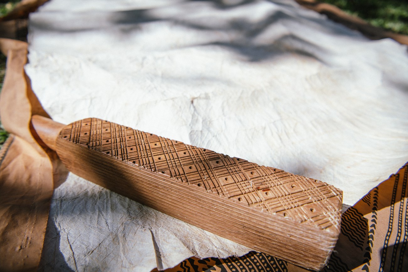 Rectangular wooden tool with diagonal lines carved in surface sits on barkcloth with imprint of design.