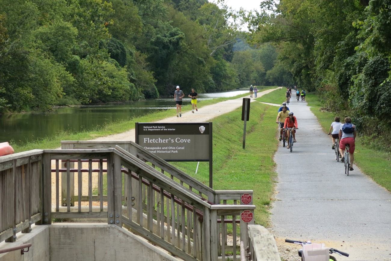 Color photograph showing two parallel trails along the water-filled canal. People walking on a gravel trail is on the far left, while people riding bicycles on a paved path is at right. Steps associated with a wooden footbridge extend over the canal
