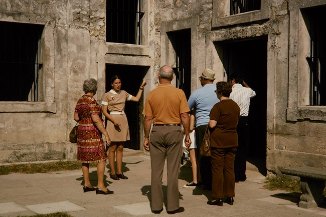 Woman in beige dress directs visitors in front of a stone building