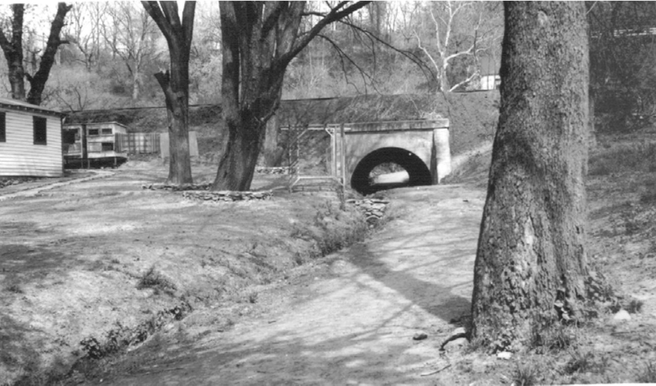 Black and white photo of the south embankment of the Chesapeake and Ohio Canal. Part of a house is visible on the left with a chicken coop behind it. In the center of the photo is the Fletcher’s Road culvert under the canal.