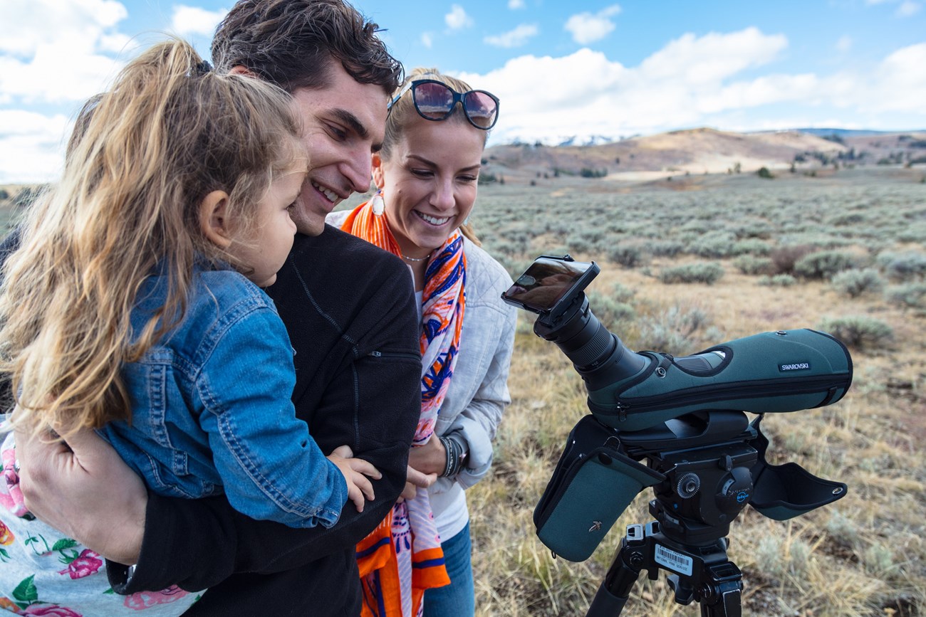 a smiling woman and a smiling man holding a child look into a spotting scope