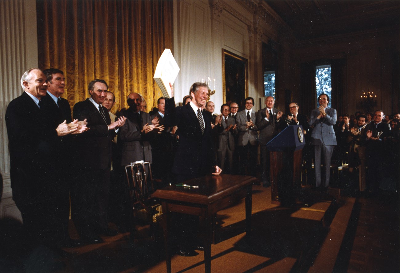 Former President Jimmy Carter stands before a desk waving a stack of papers. People surrounding him cheer and clap.