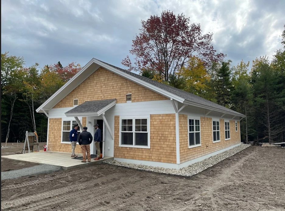 Prototype house with three people standing around front door with gravel in front and deciduous tress visible in the background