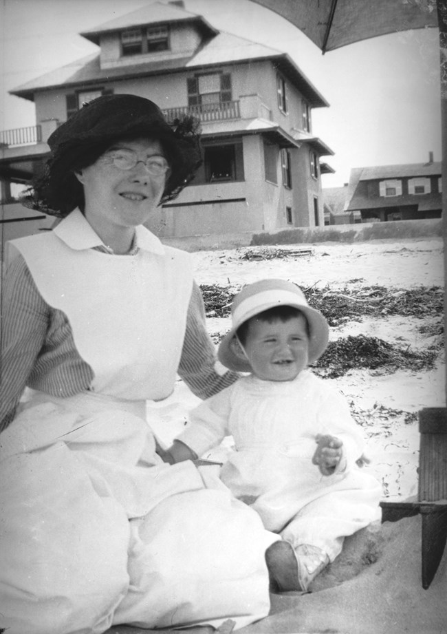 A nursemaid (left) sits with a toddler in the sand. Behind them, sand, seaweed, and several cottages are visible.