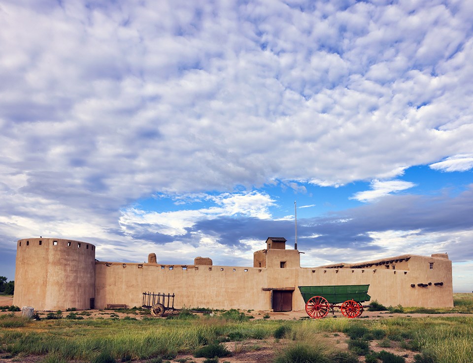 An old fort and a couple of old wagons under a cloud-filled sky.
