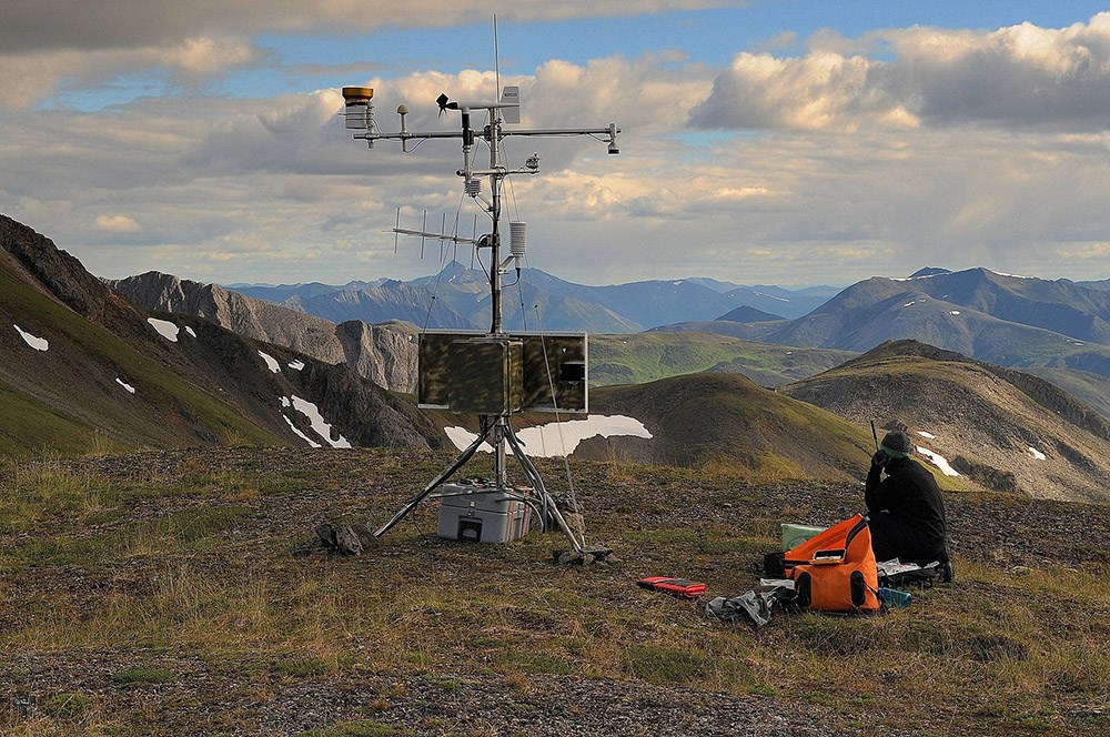 a person sits next to equipment for monitoring climate on the top of a mountain with mountains in the distance