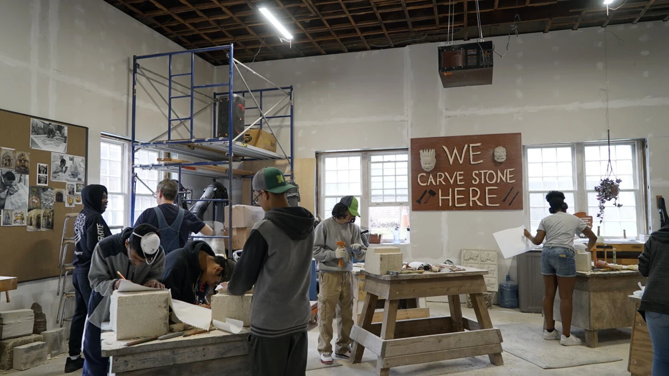 Students stand at work tables and carve large blocks of stone