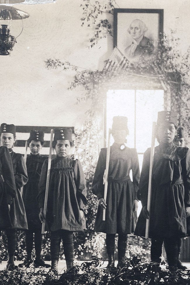 Indigenous girls in uniforms stand on stage, portraits of George Washington above them.