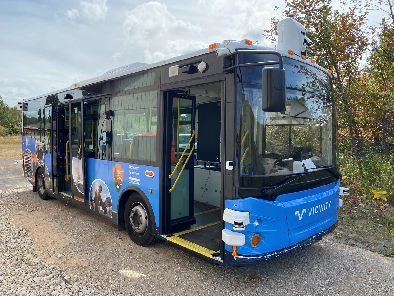 Blue and black electric bus is parked in front of small trees in a gravel parking lot