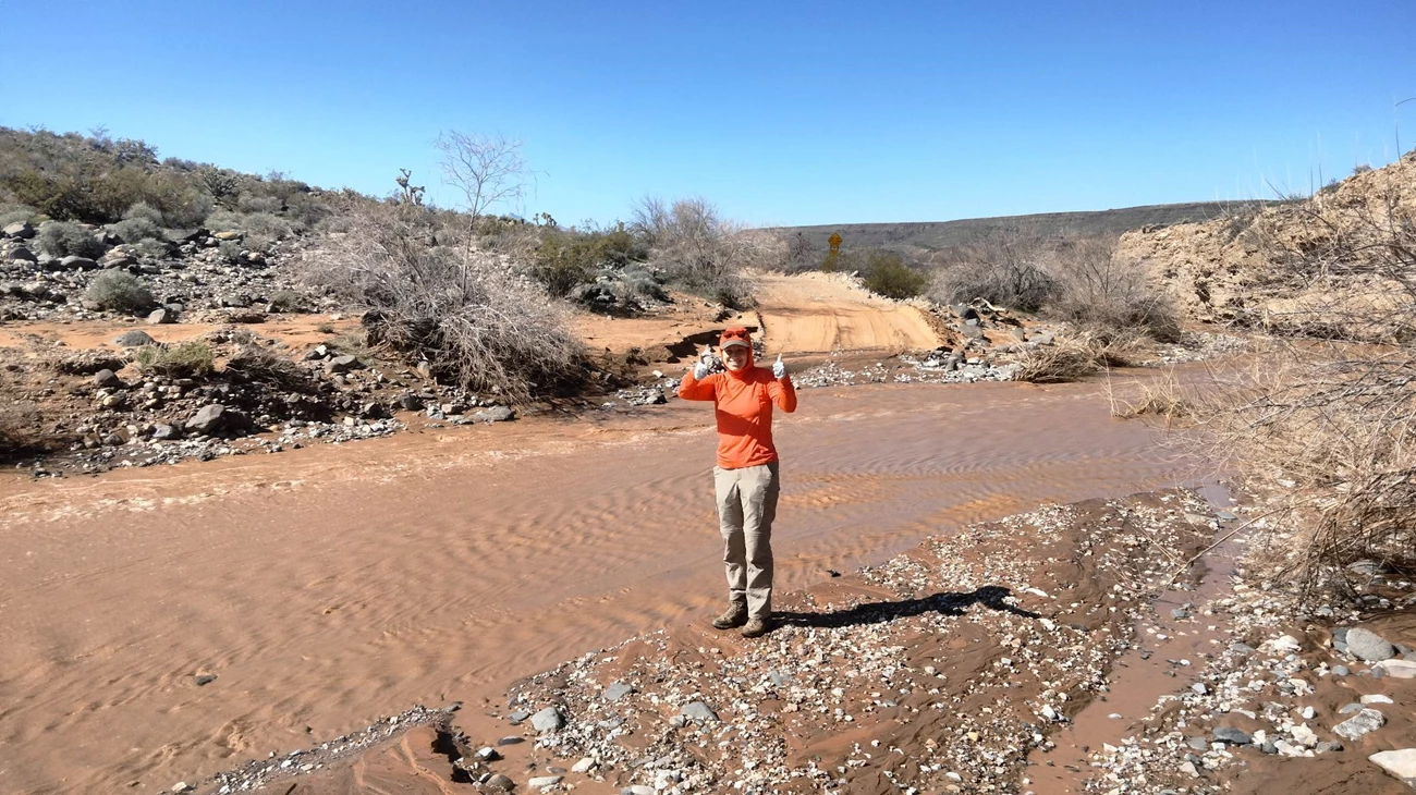 Woman gives thumbs up as she stands next to a wash flowing with muddy water.