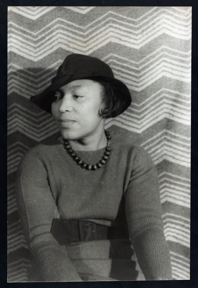 A Black woman sits looking to the left wearing a hat, beaded necklace, and sweater.