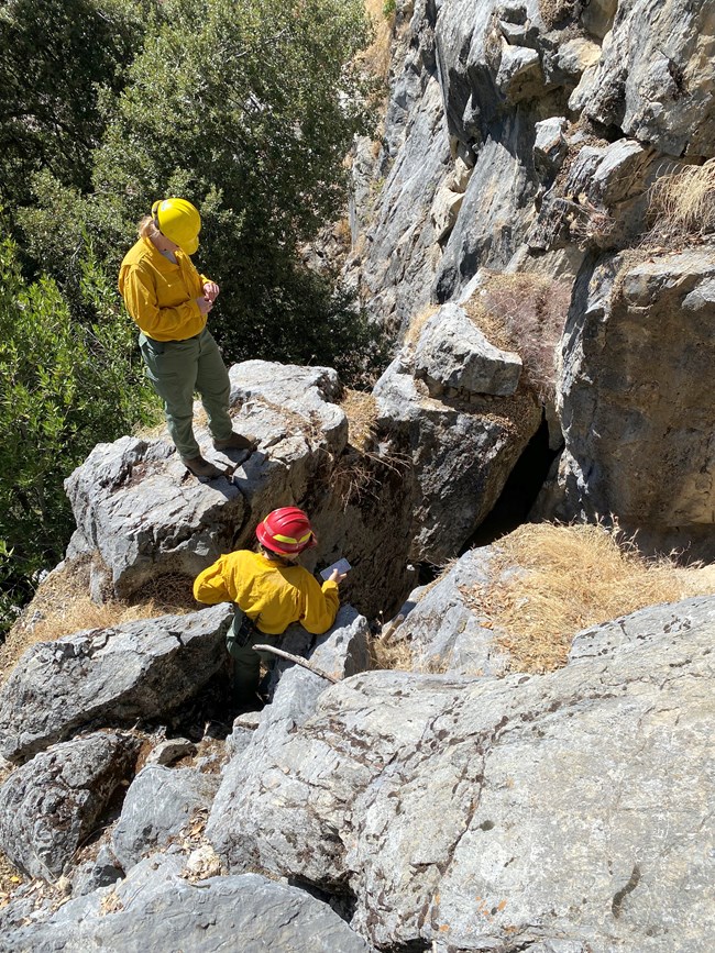 Two people in firefighting gear stand amongst large boulders near a dark opening in front of them.