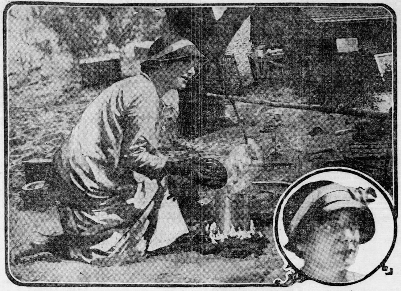 Black and white photograph of a woman wearing a long, light-colored garment and a hat. She is bent over a campfire, tending to a pot on the flames. Sand, trees, and wooden structures are seen in the background.