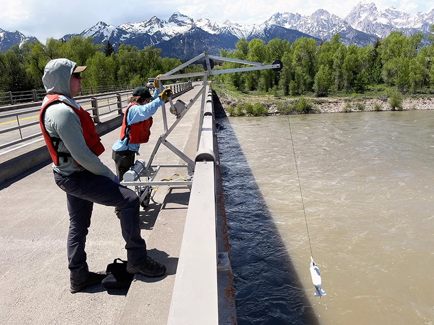 Two scientists standing on a bridge over a river turning a crank on a crane to lower a reel with equipment for collecting water samples into the river.