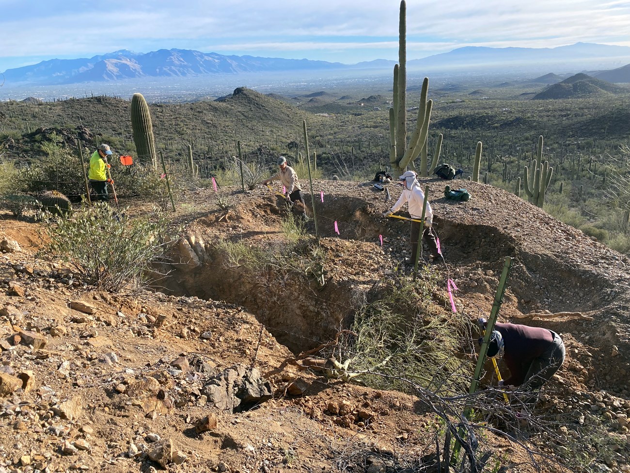 A crew of workers using tools to fill a large opening of an abandoned mine site. There are mountains and cacti in the landscape.