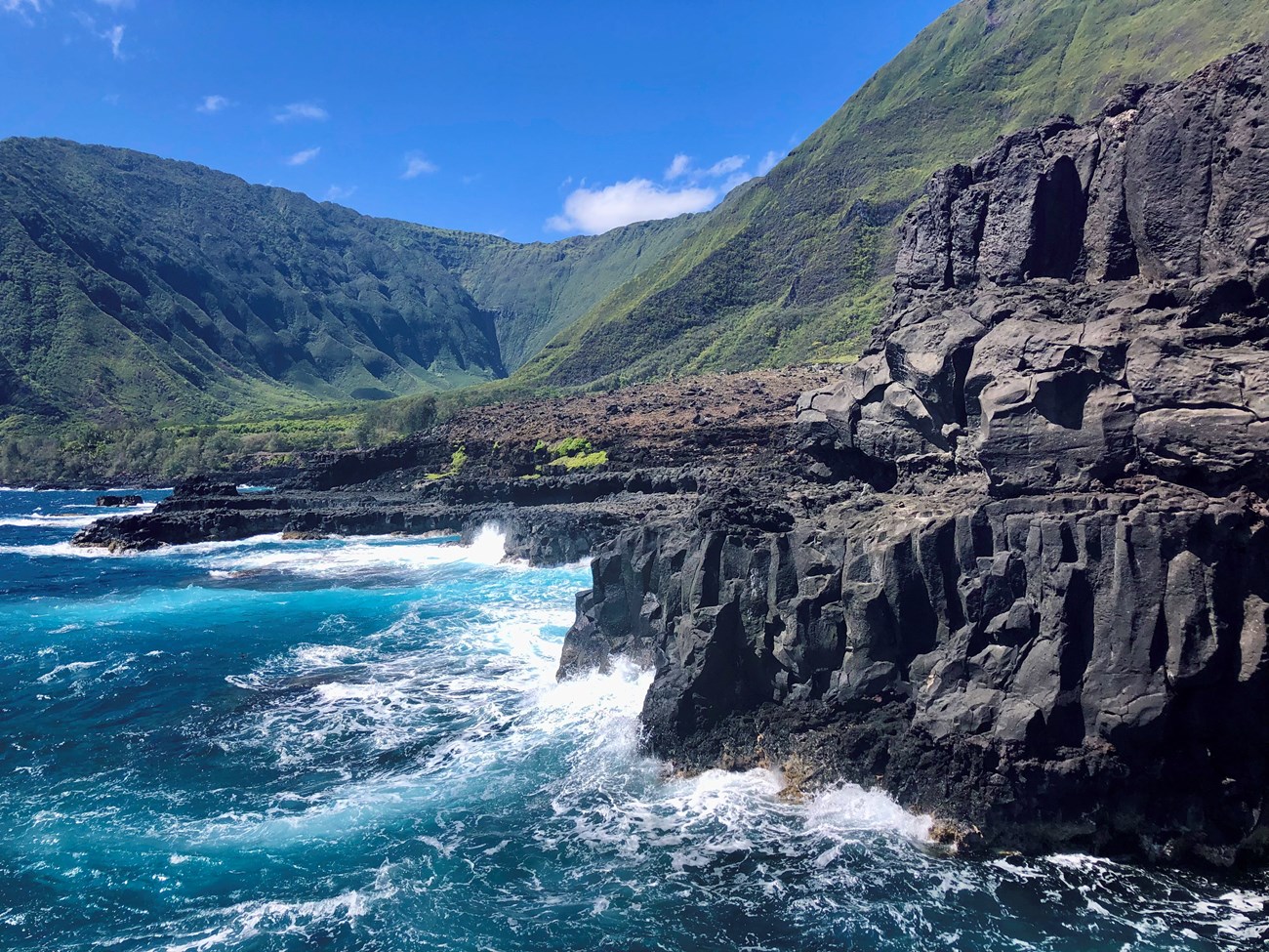 Turquoise waves crash against volcanic coastline, deep, lush valley and mountains in background.
