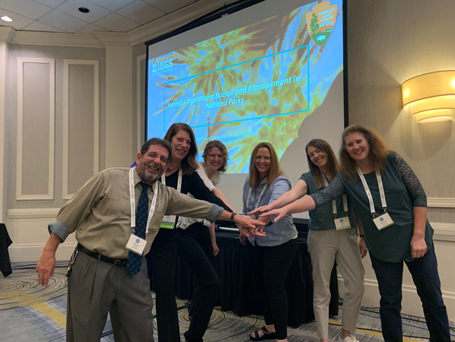a group of six presenters extend their arms and hands into the middle of their semi circle in a "go team" gesture in front of a projection screen with a title slide that says "harmful algal blooms science and management in national parks"