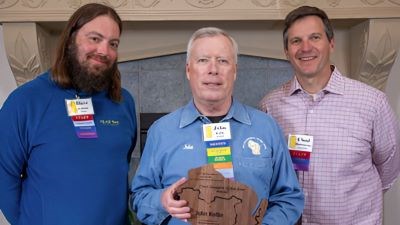 A man stands holding an award, on either side of him stands two men smiling.