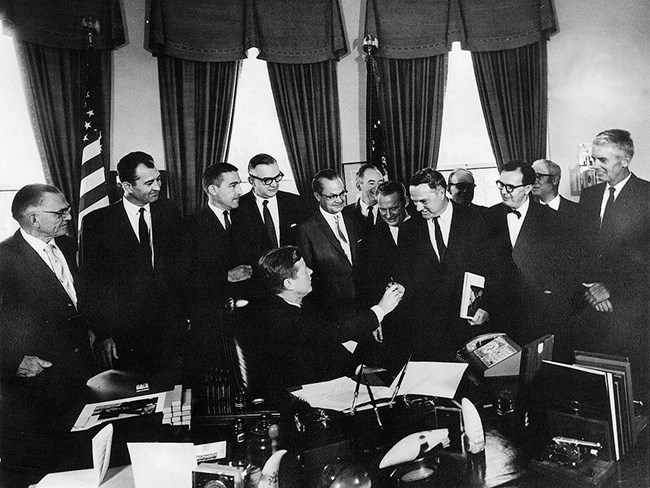 In an ornate office, eleven men dressed in suits look on from behind a large desk as a man seated at the desk turns in his chair to hand a pen to a man standing to the left of him.