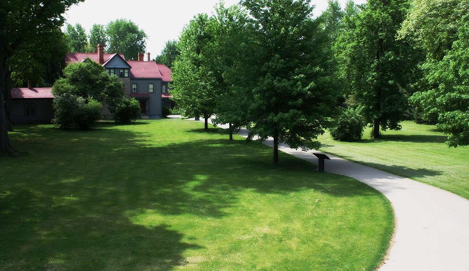A walkway lined with leafy trees leading to a large house with tall chimneys