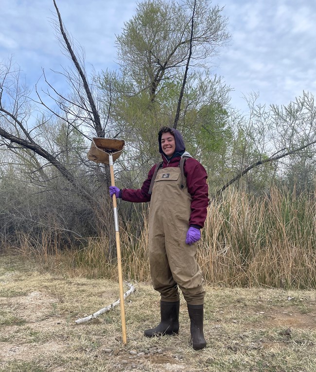 Woman wearing waders and holding a net for collecting stream invertebrates.