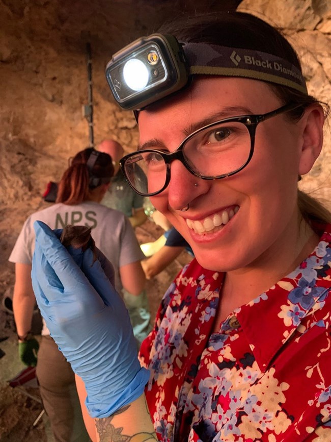 Woman wearing headlamp holding a bat with gloved hand and smiling at camera.