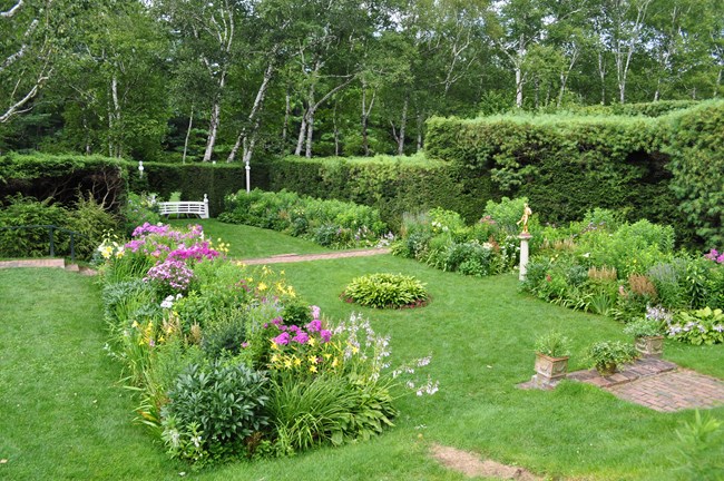 Short grass grows between rectangular planting beds with blooming perennial flowers, with brick walks and sculptures. It is surrounded by a tall hedge.