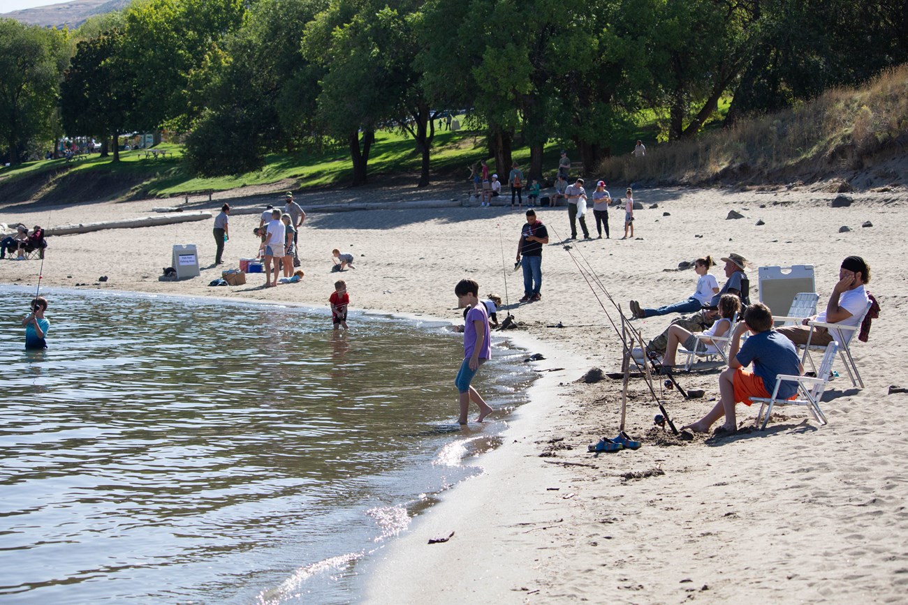 A number of children and adults spread out on a sandy beach. Many children are holding fishing poles and fishing from the shore.