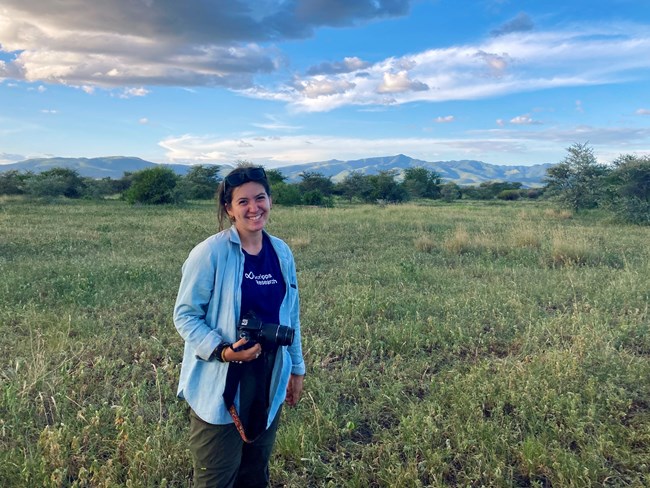 Woman stands in field smiling and holding a camera.