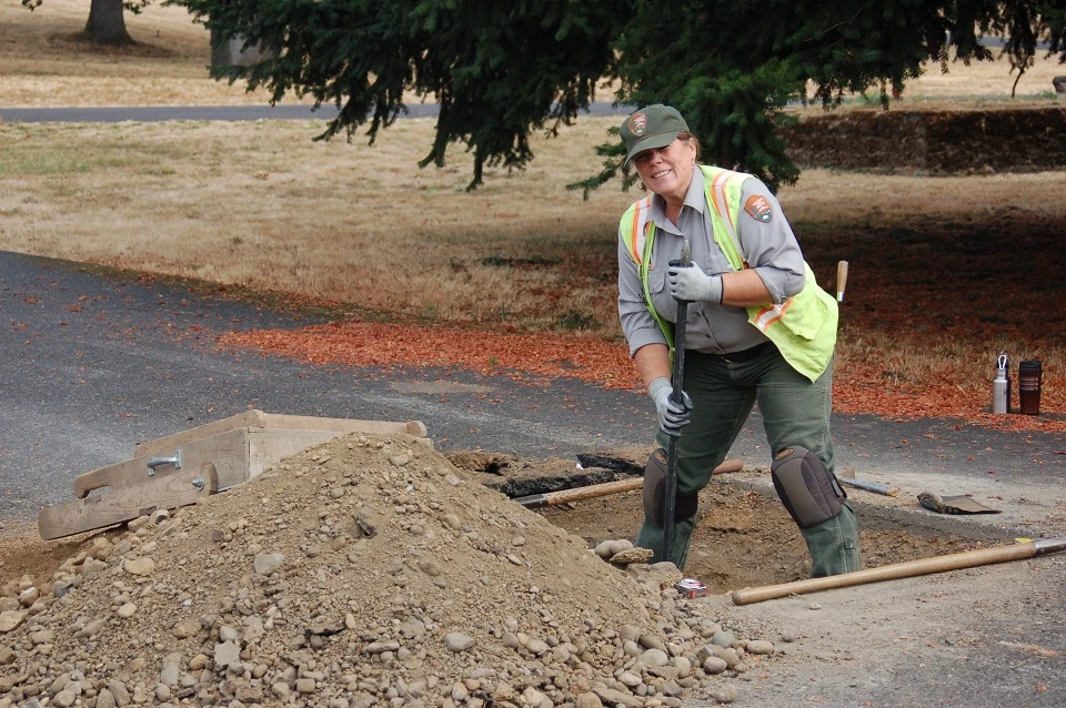 A woman wearing a National Park Service uniform stands in an excavation unit holding a shovel.