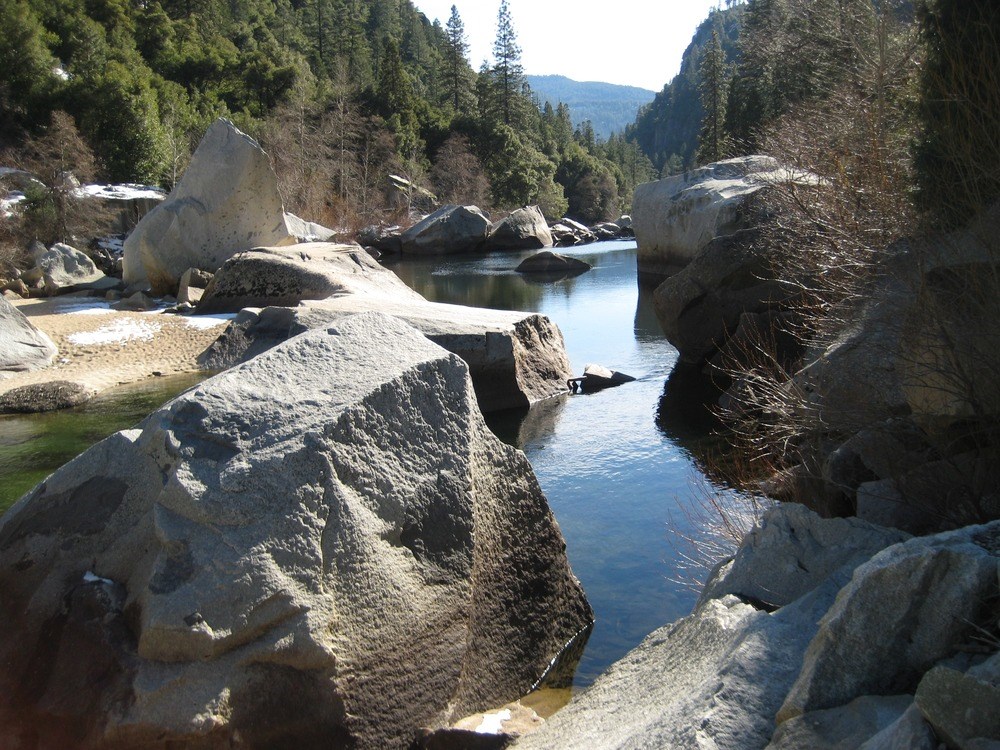 A view through boulders of the Merced River with mountains in the boackground.