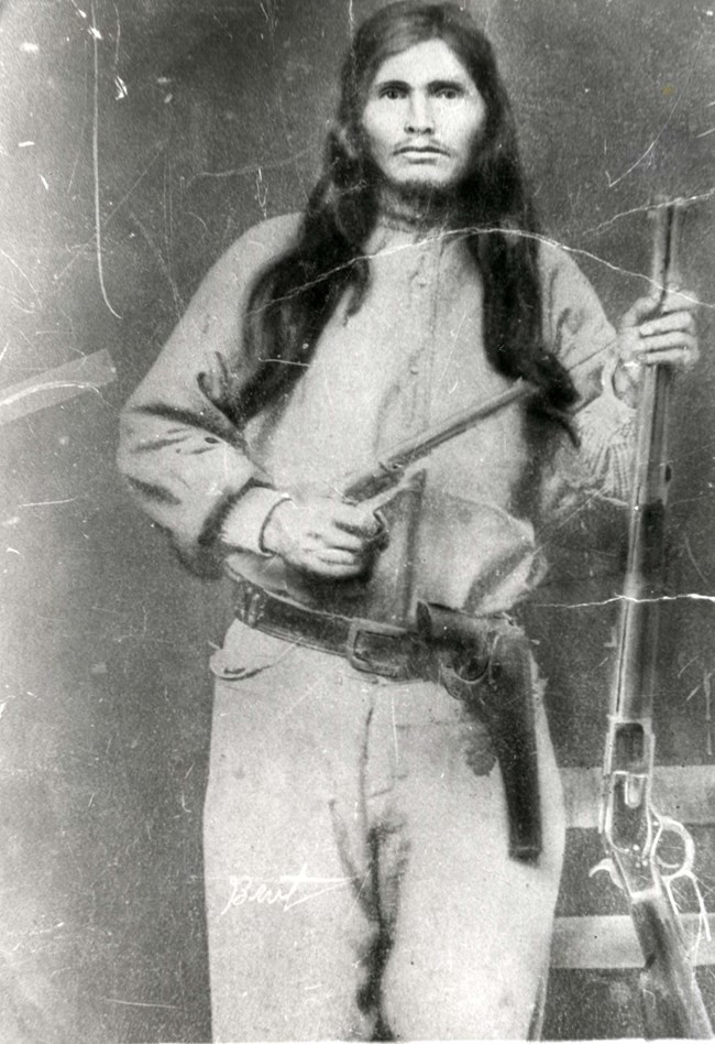 Historic photo of Ned Christie, a man with long dark hair standing holding two guns with a serious expression.