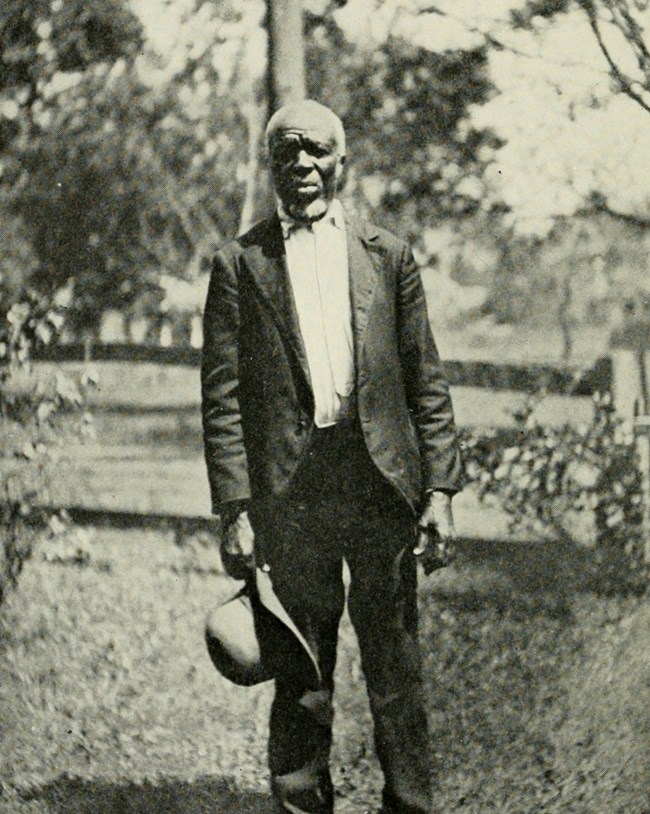 A Black man stands in a suit outside holding his hat in his hand.