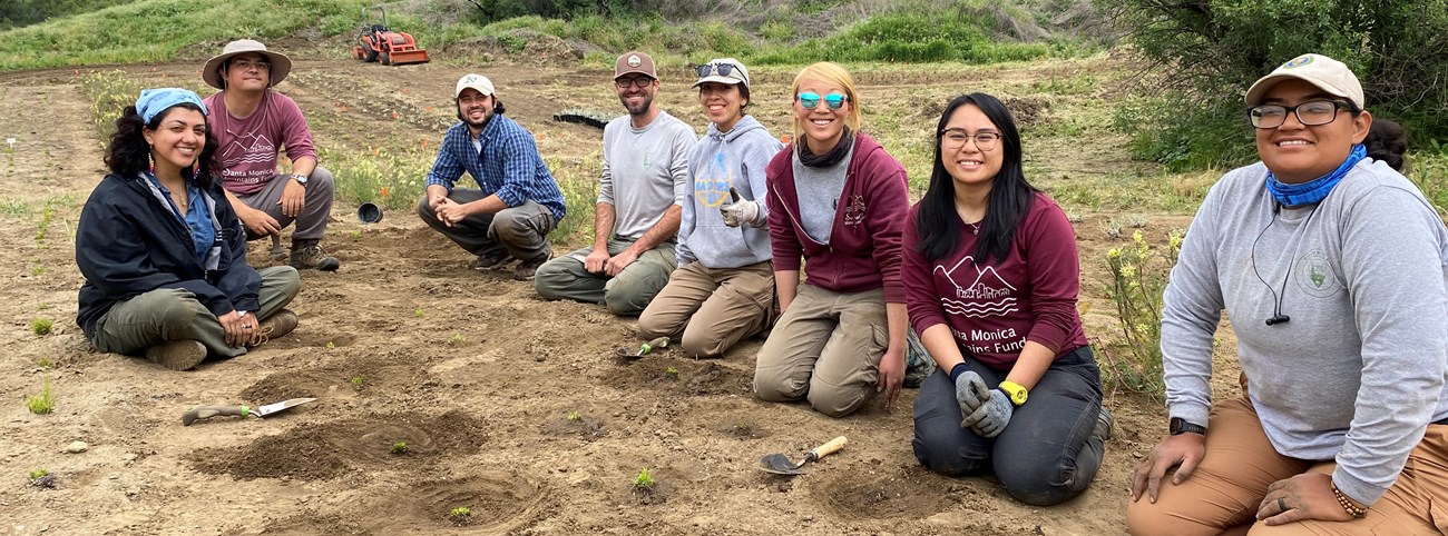 A diverse group of people sit and kneel on the ground in middle of field with small plants.