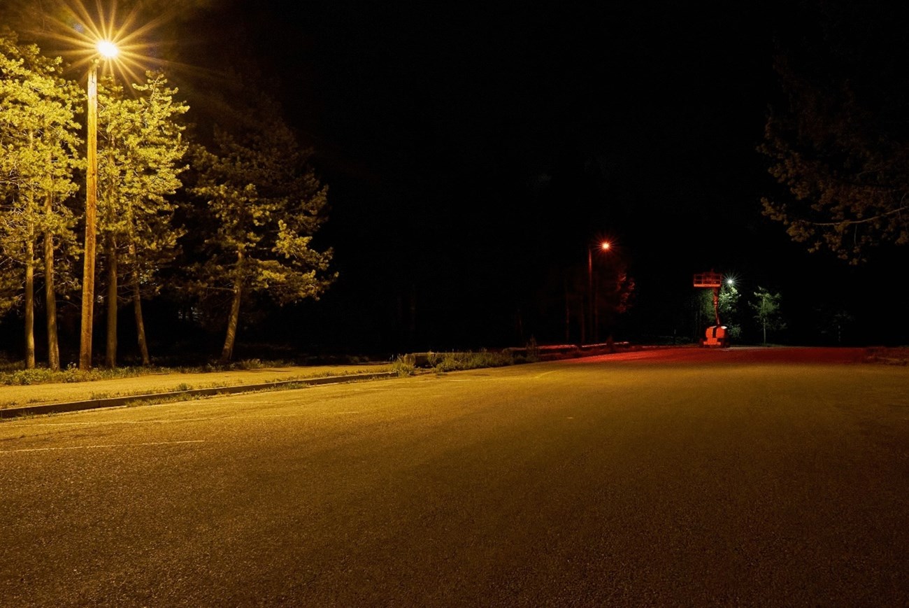 Looking down an empty parking lot at night lined with streetlights of different colors. There's a bright, yellowish standard light in the front left, then a red light, and then a cool white light in the distance.