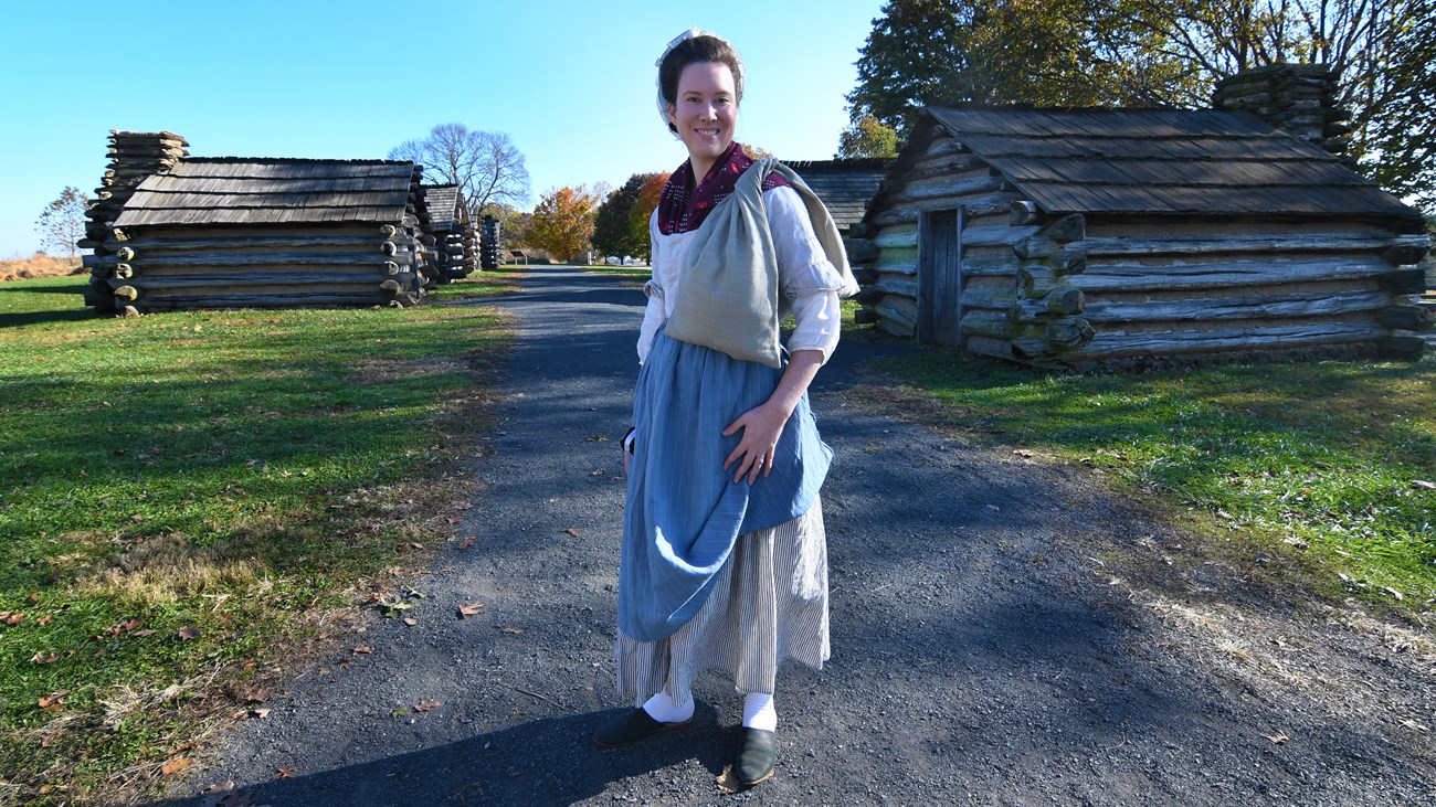 photograph, outdoors, smiling woman, 18th century clothing, market wallet, log huts