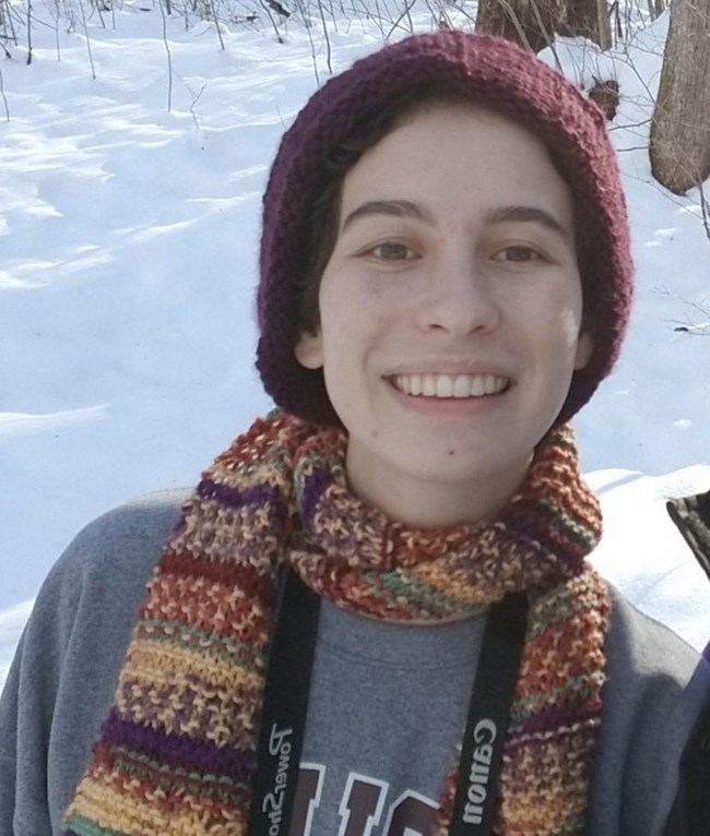 Woman smiling and wearing winter hat and scarf, with snowy forest scene behind her.