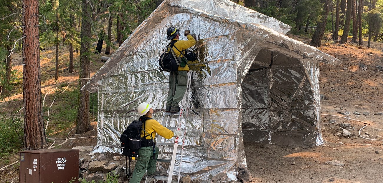A person in wildland firefighting personal protective equipment (PPE) stands on a ladder putting foil on a structure while another holds the ladder below.